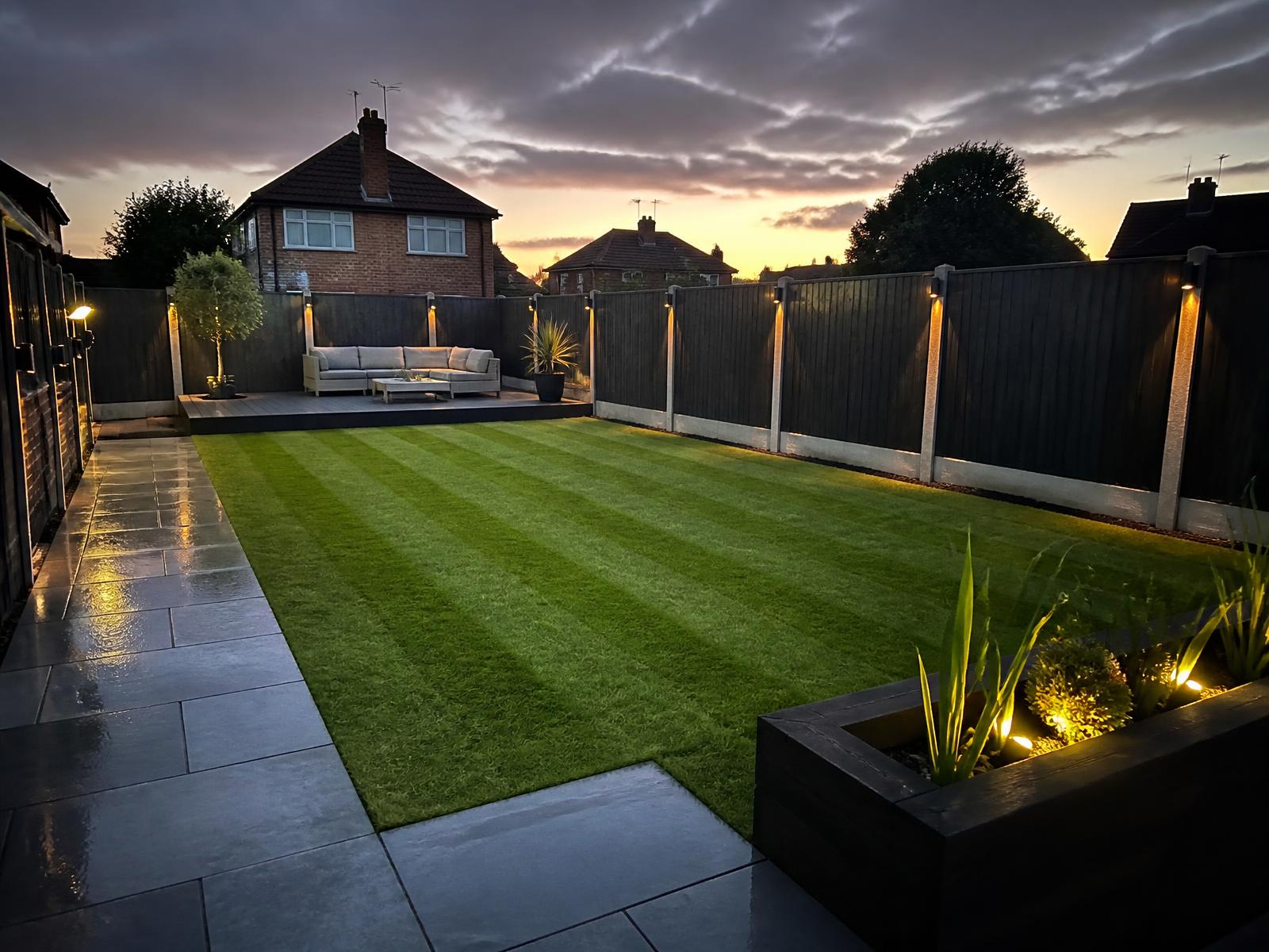 Finished garden with striped lawn, dark fencing and paved path at dusk